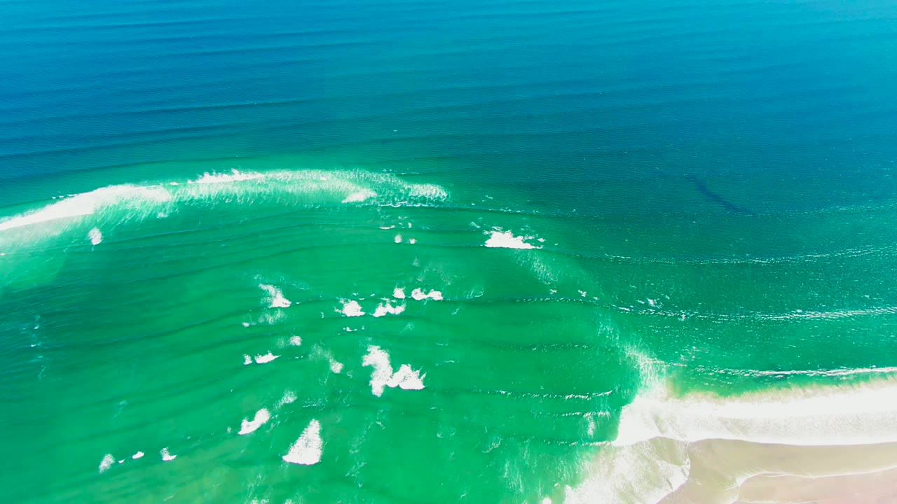 White foam bands break across turquoise water beside a sandy edge.