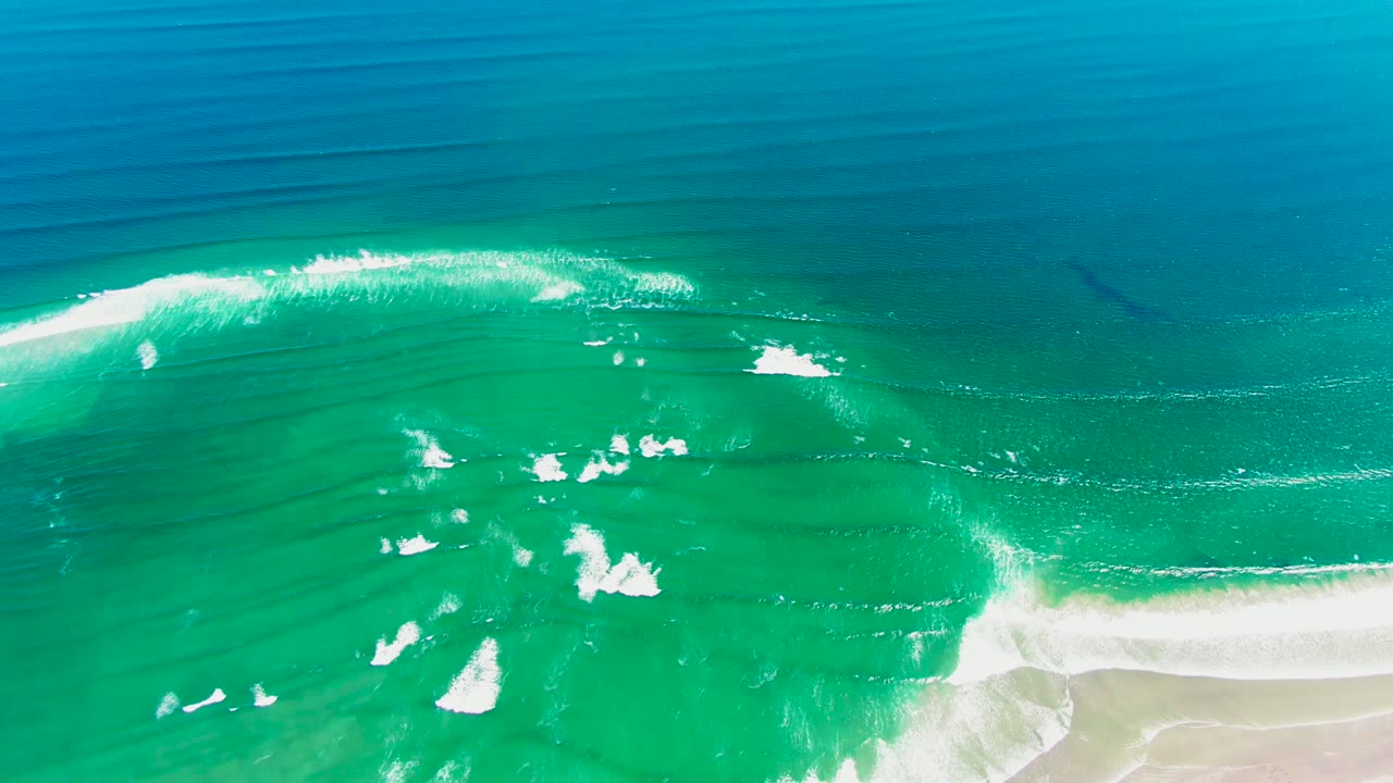 Aerial frame from La Bocana, Baja California Sur showing deep blue water across the top, turquoise shallows below, foam streaks through the middle, pale beach at lower right, and no visible people.
