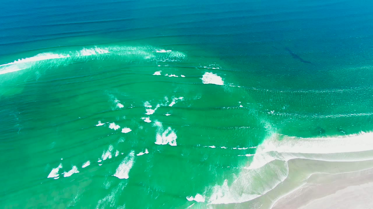 Overhead view of turquoise ocean waves and white foam approaching pale sand at La Bocana, Baja California Sur, with no visible people.