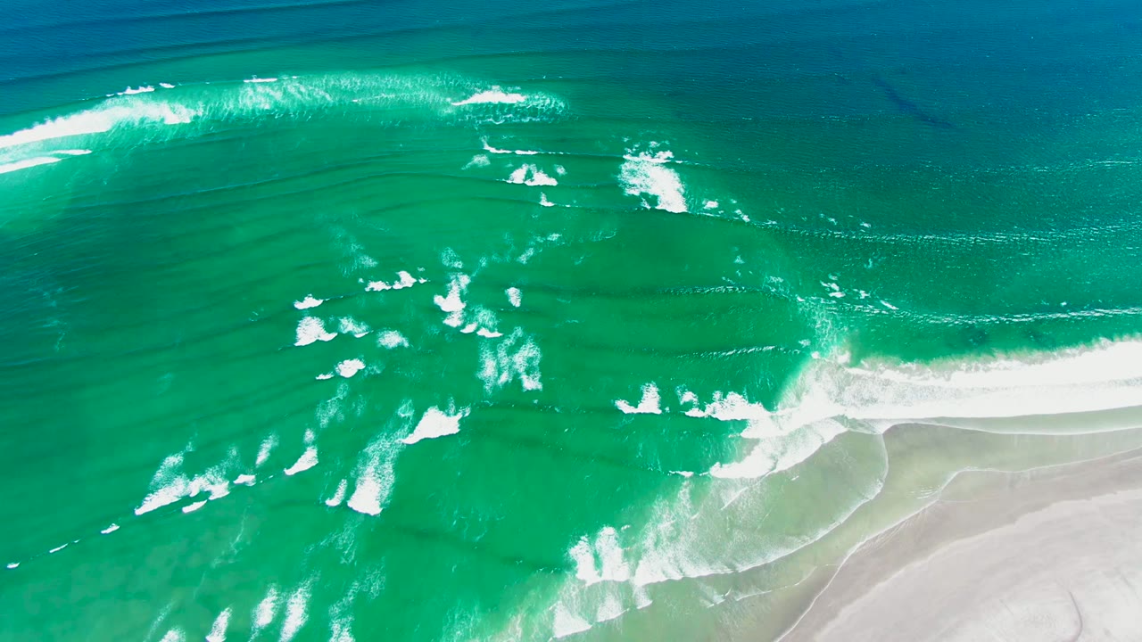Overhead view of turquoise ocean and white foam rolling toward pale sand in La Bocana, Baja California Sur, with no visible people.