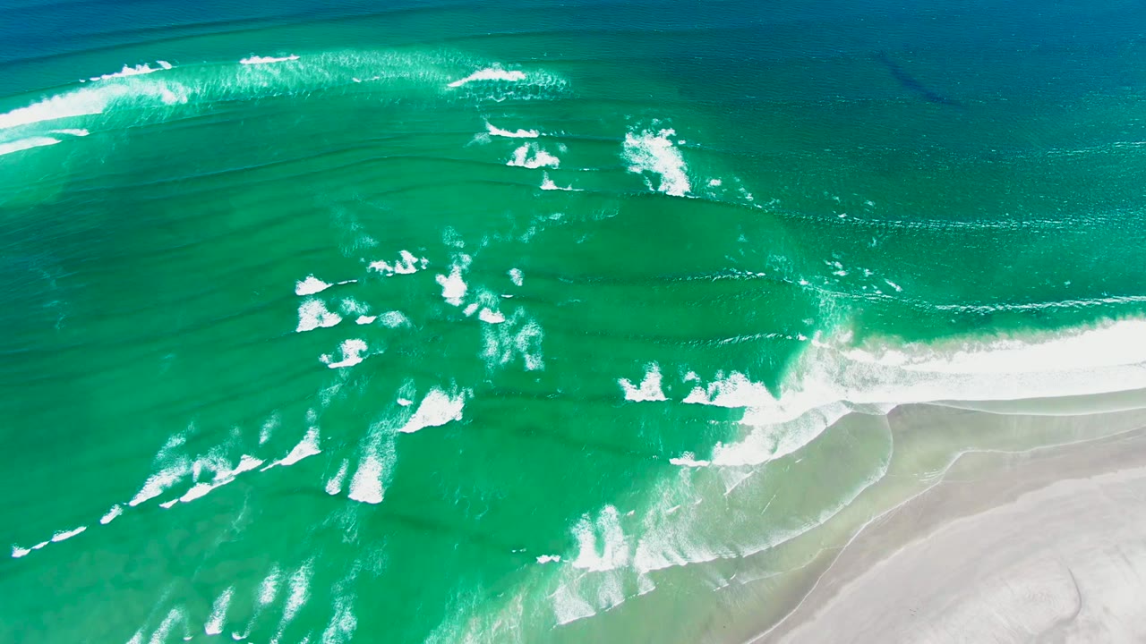 Drone-height overhead view of surf curling across turquoise water toward the bright sandy shoreline at La Bocana, with no visible people.