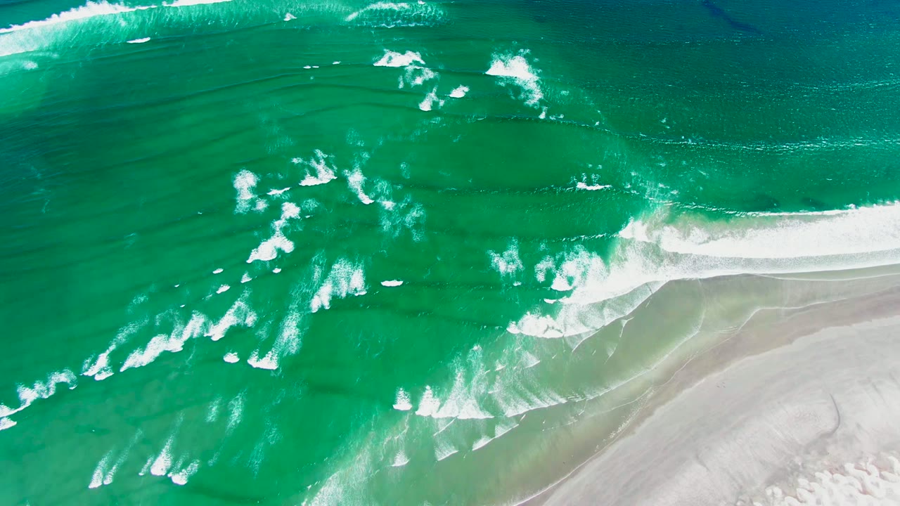 Top-down aerial frame of turquoise waves and white foam approaching pale sand at La Bocana, Baja California Sur, with no visible people.
