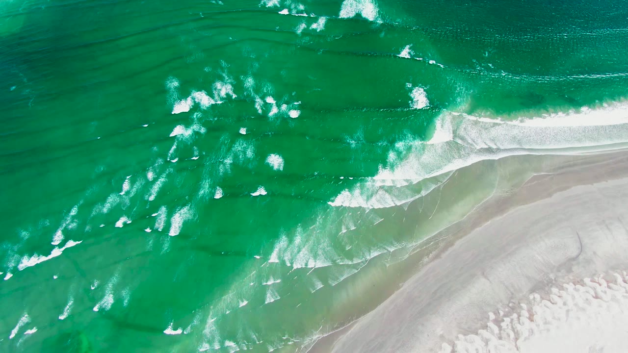 Overhead La Bocana surf view with small white breakers scattered across green-blue water and pale sand curving along the lower right, with no visible people.