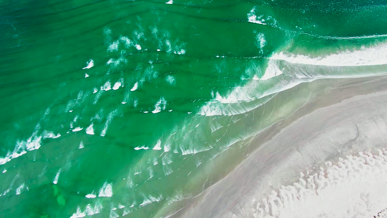Top-down aerial frame from La Bocana, Baja California Sur showing emerald water, diagonal white surf lines, and pale sand at the lower right with no visible people.