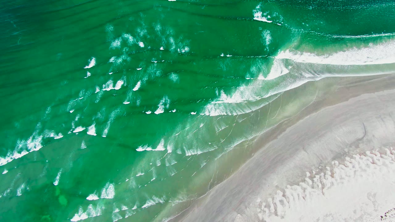 White foam lines spread across green water beside a sandy shoreline
