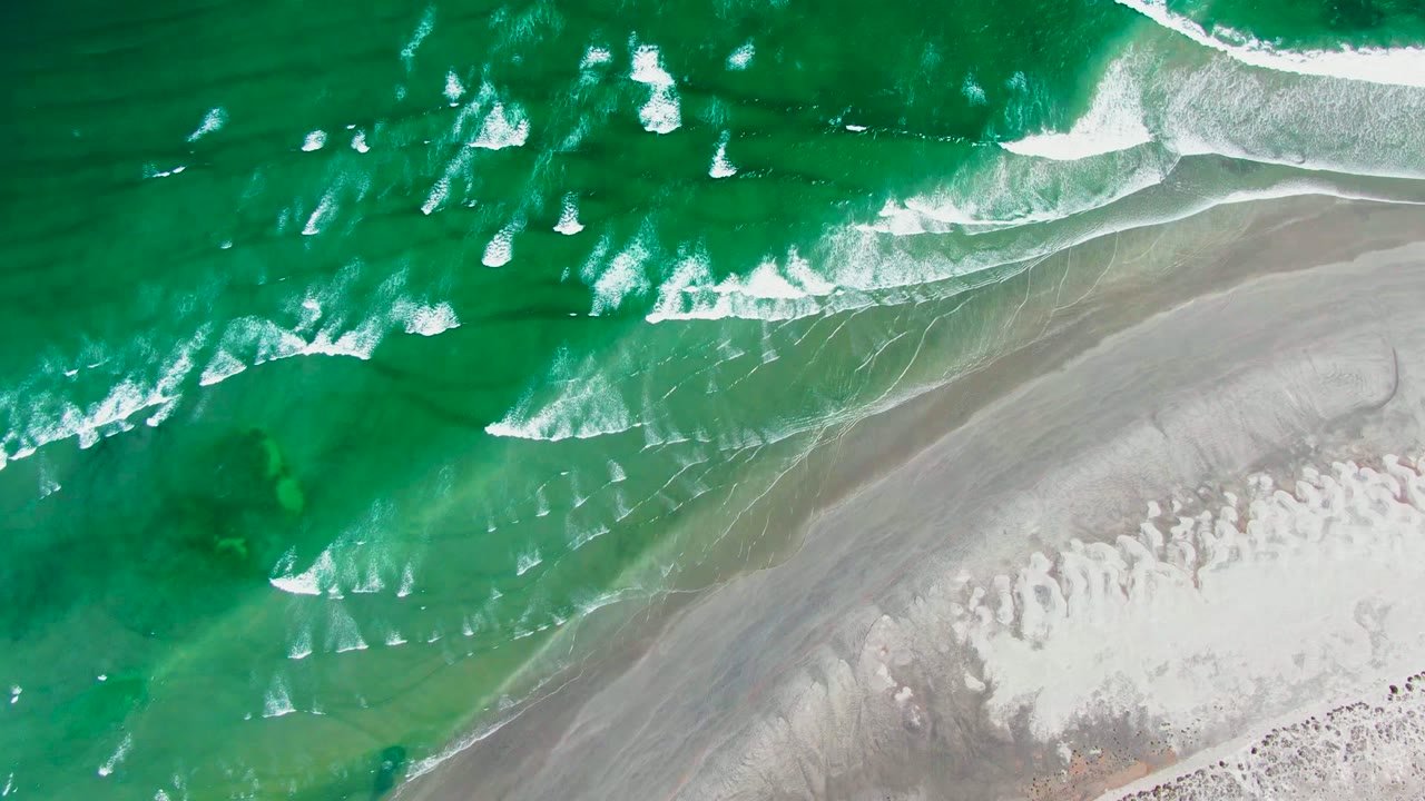 Overhead view of green surf and white foam approaching gray wet sand and pale beach at La Bocana, Baja California Sur, with no visible people.