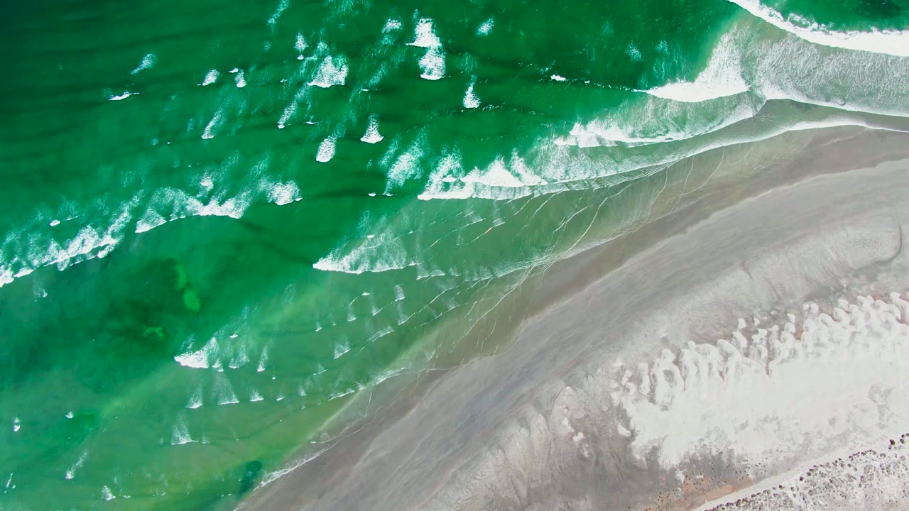White foam lines spread through green water near a sandy shoreline.