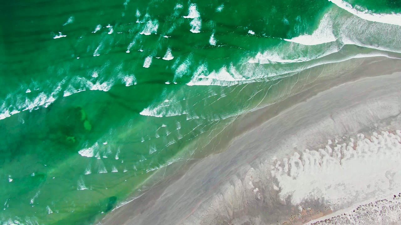 Aerial view of diagonal white surf breaks beside a gray and pale sandy beach in La Bocana, Baja California Sur, with no visible people.