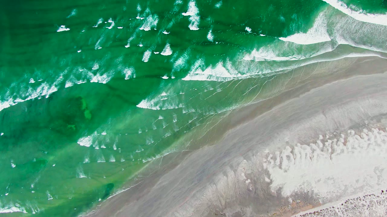 Green waves and foam move over shallow water toward empty sand.