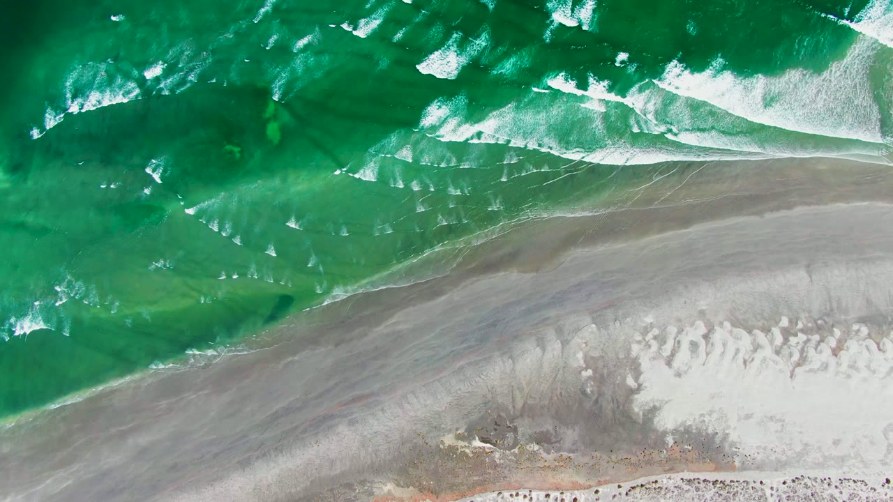 Aerial view of green surf and white foam washing toward pale sand in La Bocana, Baja California Sur, with no visible people.