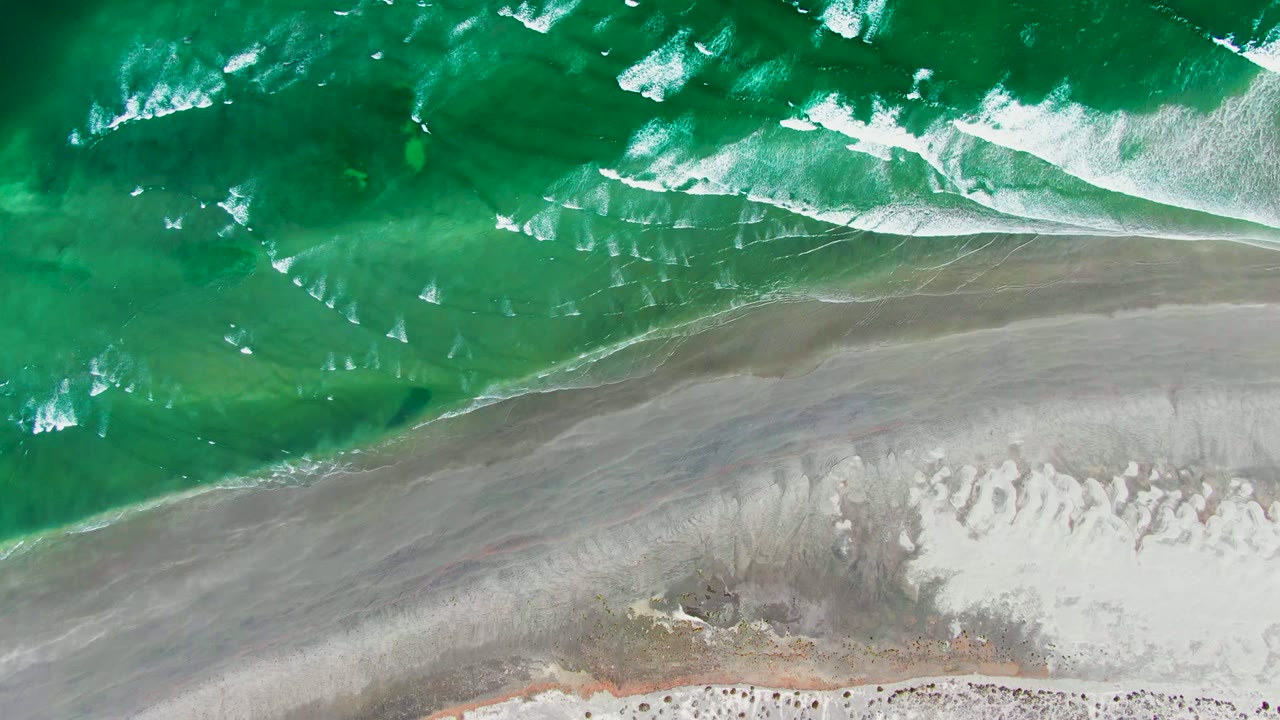 Top-down La Bocana beach frame with green ocean across the upper half, pale sand below, and broken white foam along the edge, with no visible people.