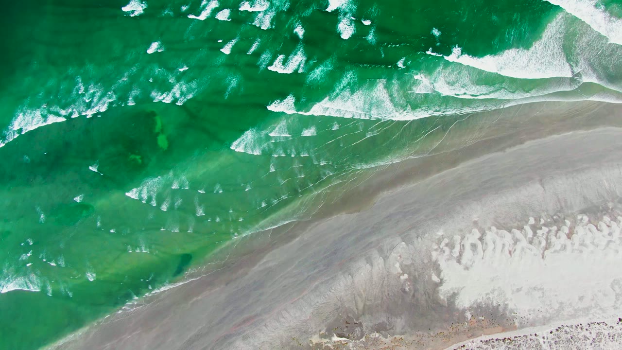 Top-down view of white surf bands over green water with a broad strip of gray wet sand and lighter dry sand at La Bocana, no visible people.