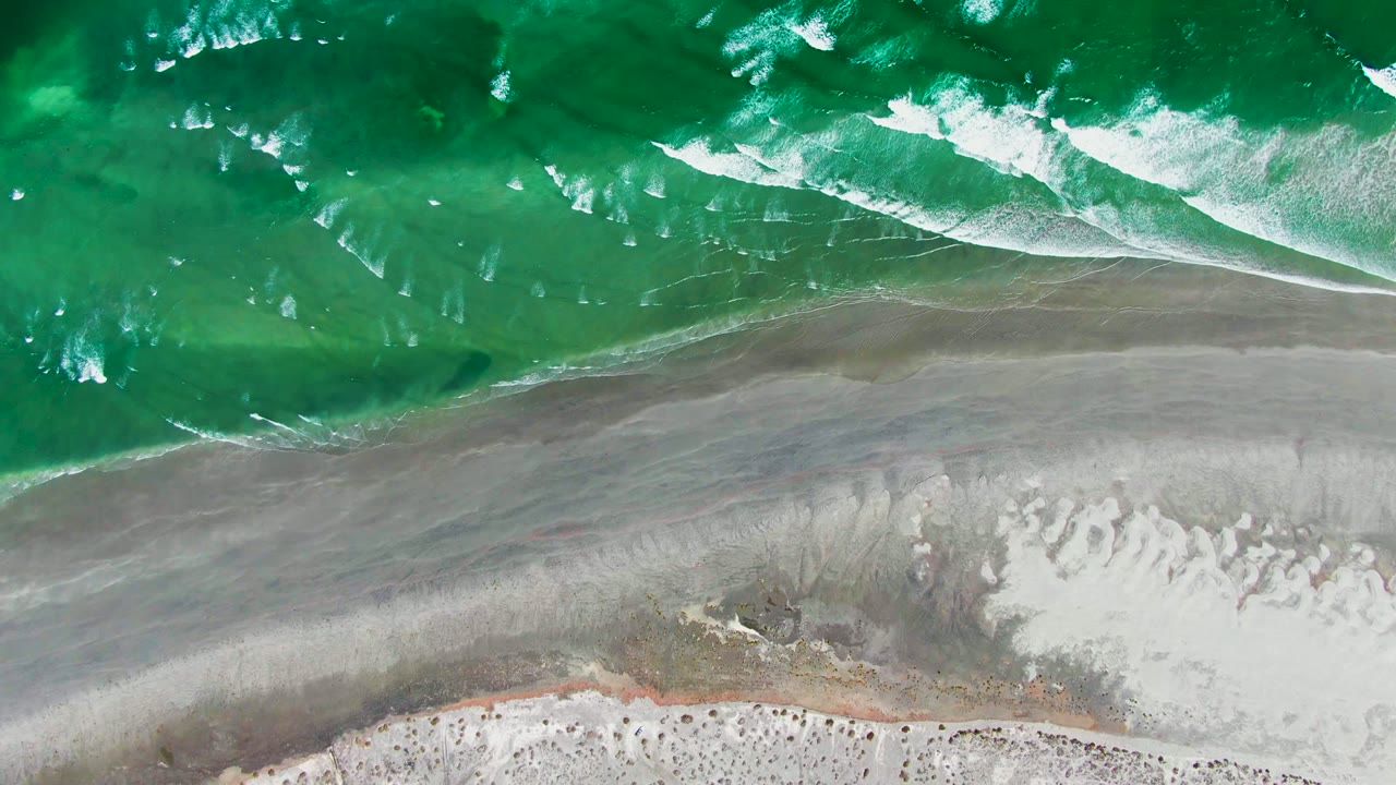 Overhead view of green ocean water and white surf reaching gray wet sand on the La Bocana shoreline, with pale beach below and no visible people.