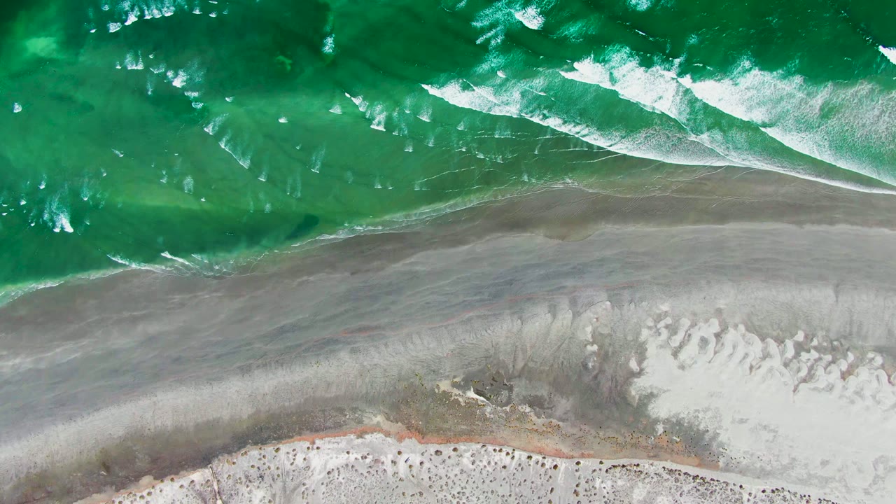A diagonal band of white foam spreads from the green water toward smooth gray sand at La Bocana, with the dry beach edge visible at the bottom and no visible people.