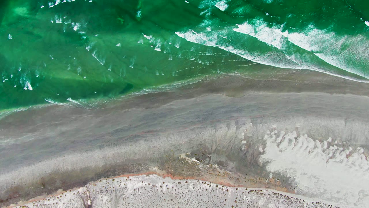 Green coastal water churns with broken white foam above a broad strip of gray wet sand on the La Bocana beach, with no people or structures visible.