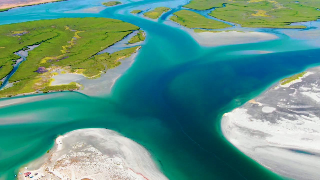Mid-clip aerial frame over La Bocana where the turquoise shallows and deep channel curve around low green islands and a bright white sandbar, with no visible people.