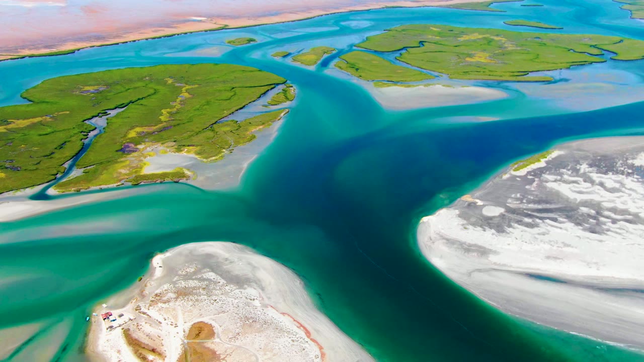 Aerial frame over La Bocana, Baja California Sur, showing turquoise channels around green islands, pale sand flats, white shoreline, and a tiny boat on the lower sand with no visible people.