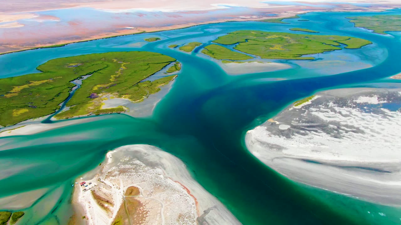 Aerial view at La Bocana showing blue-green coastal water wrapping around low green islands, bright sand flats at right and lower frame, and no visible people.