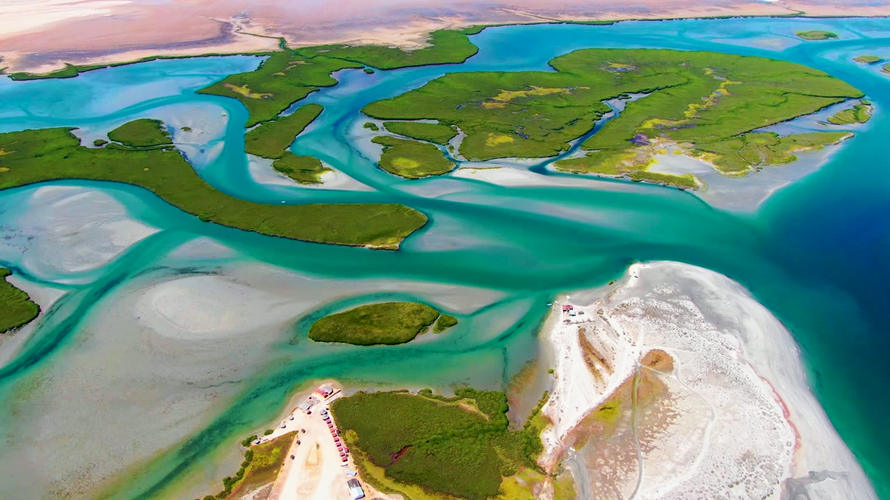 Wide aerial frame at La Bocana with shallow turquoise water wrapping around green islands, gray-white sand flats to the left, white shore at the lower right, and no visible people.