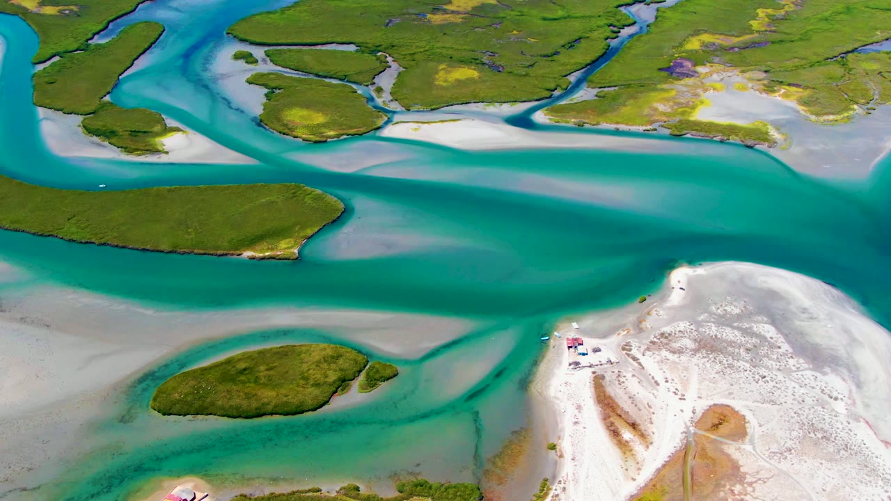 Overhead La Bocana coastal frame with deep turquoise water in the middle, green islands across the top and left, and a white sandy point at the lower right, no visible people.
