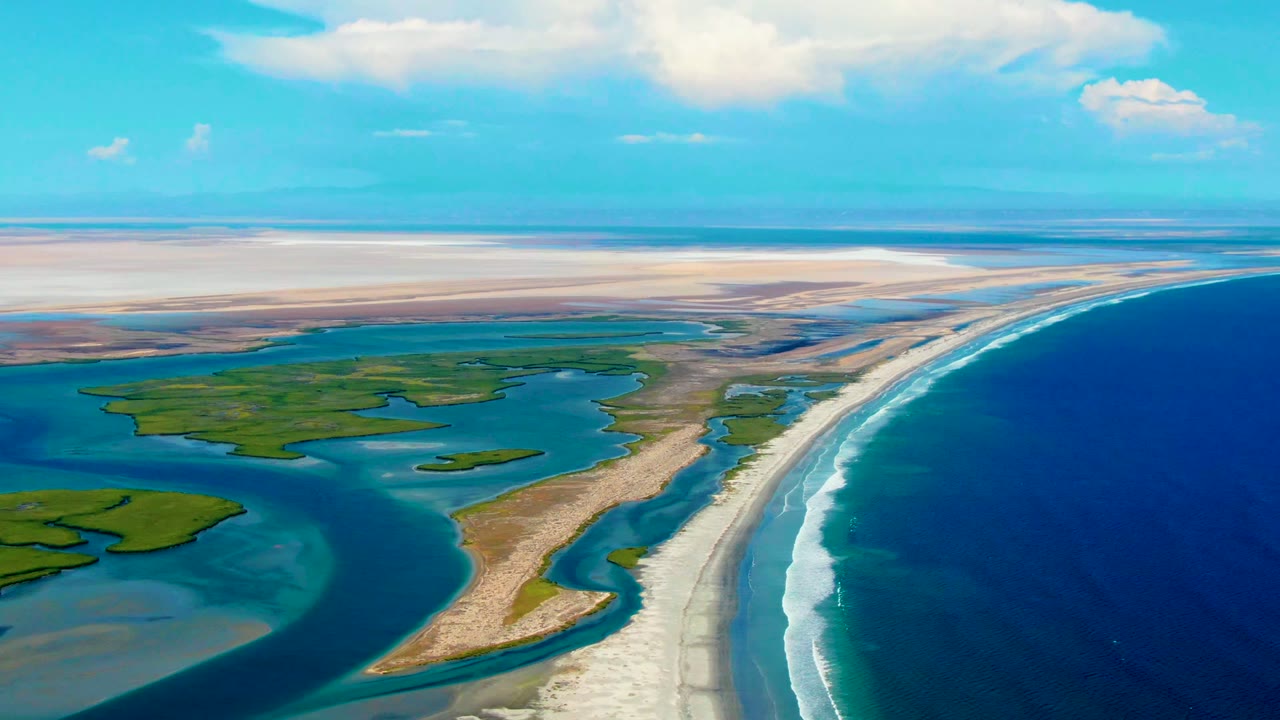 Wide aerial coastline with surf, sandbar, inland water, and green land patches.