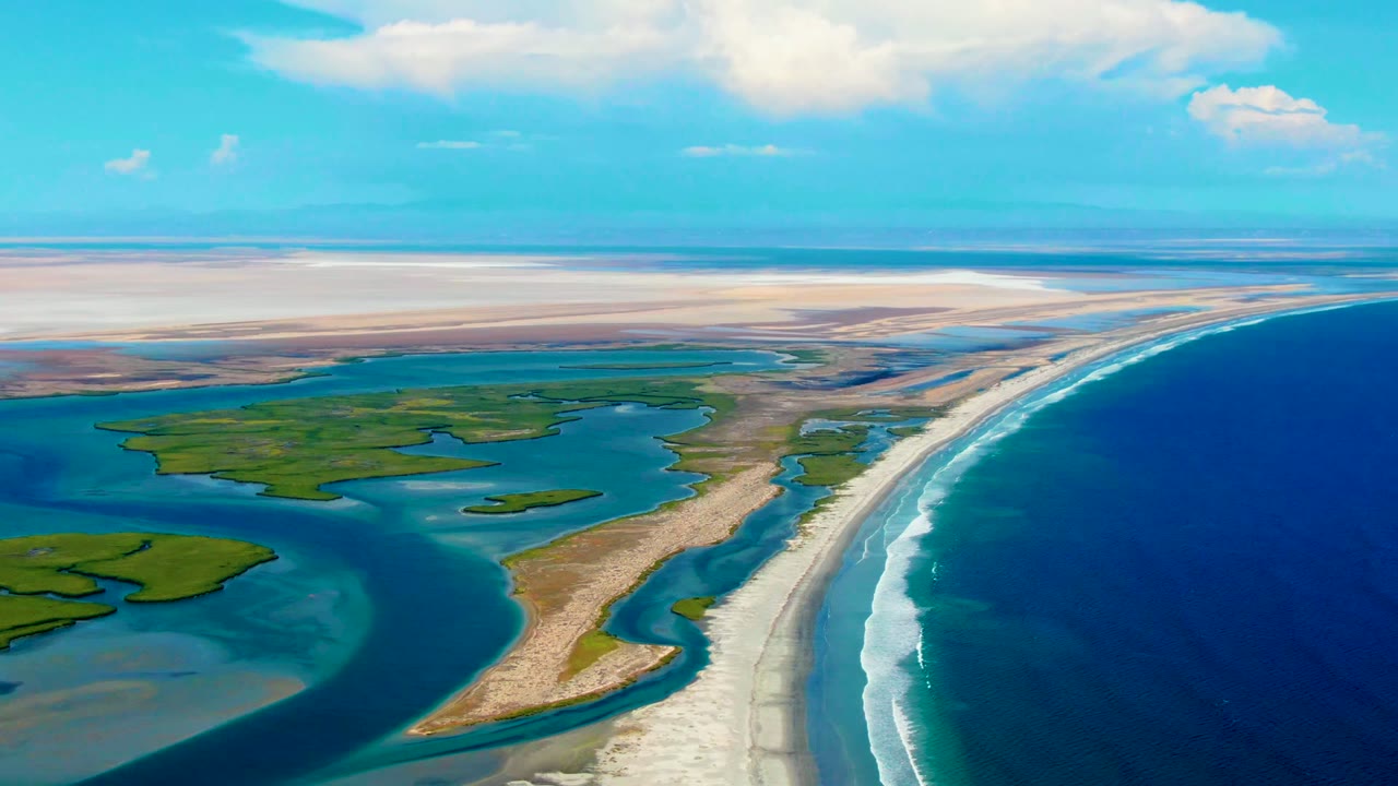 Drone-style aerial over La Bocana with the ocean filling the right side, lagoon channels and green islands on the left, pale sand across the middle, and no visible people.