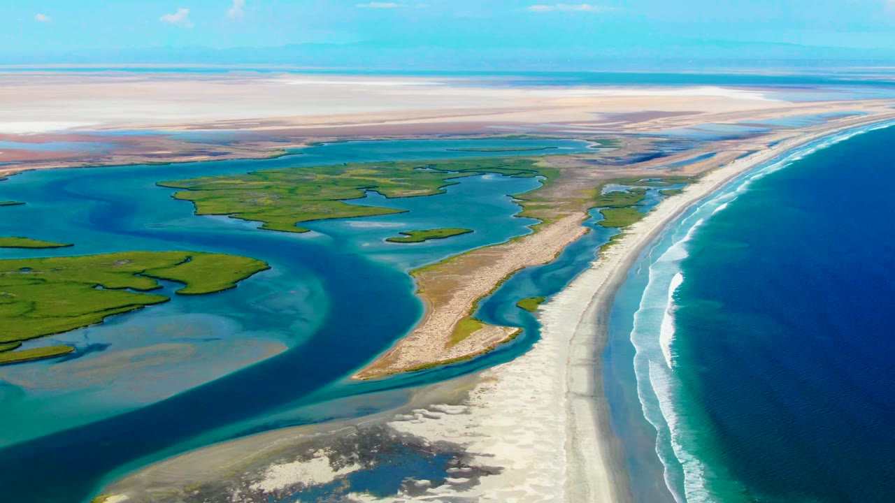 High aerial view at La Bocana, Baja California Sur, with turquoise inlet water around green marshes, a pale sand beach along the right edge, ocean surf, and no visible people.