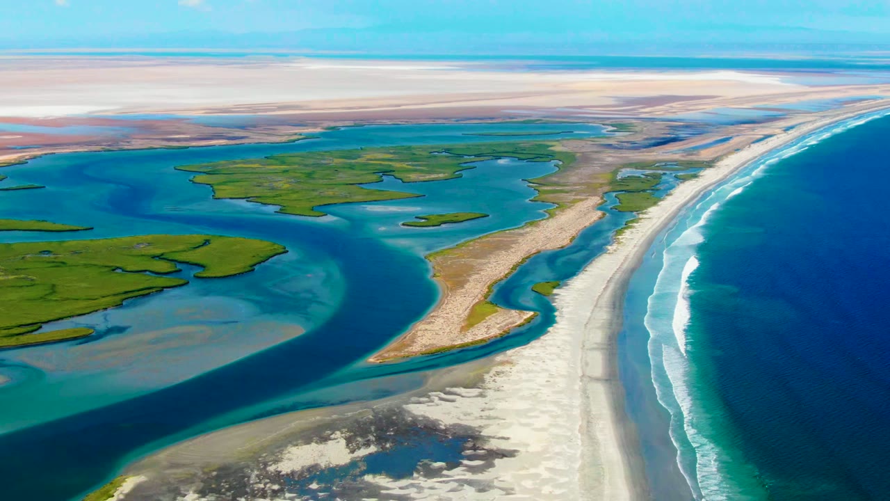 Drone view over winding blue channels beside green marsh and a long sandy shoreline.