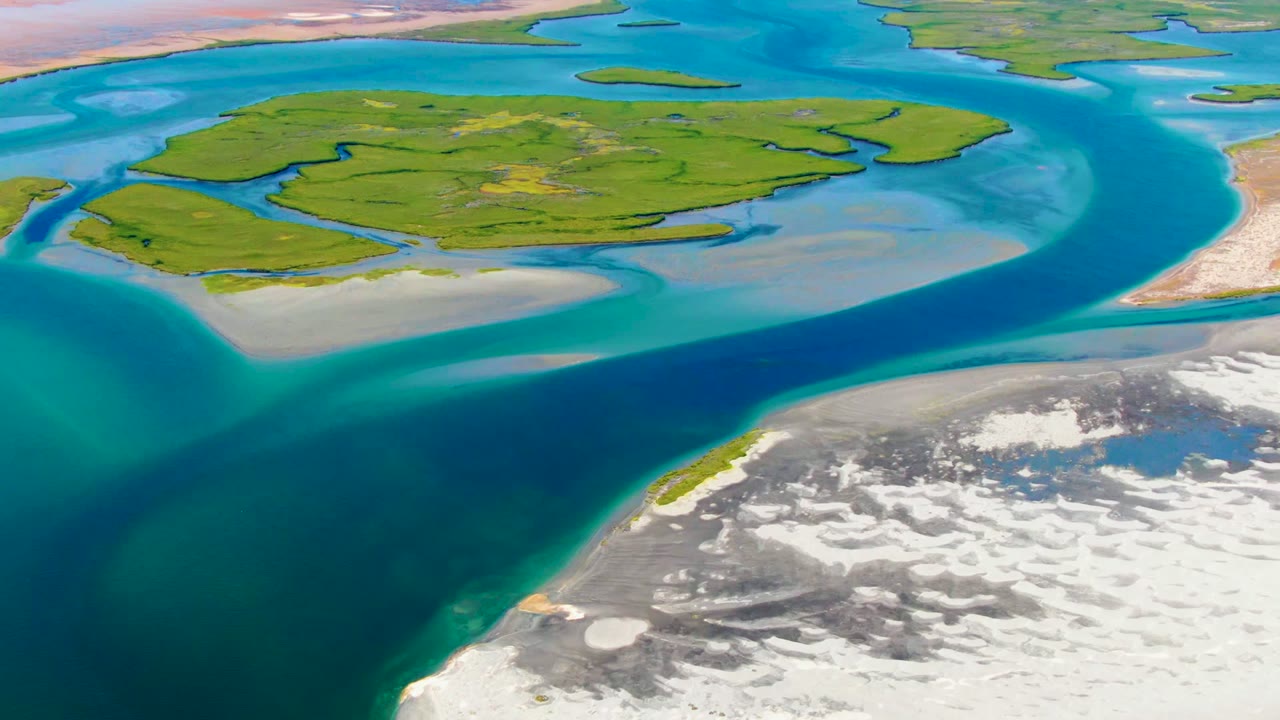Opening aerial frame over La Bocana, Baja California Sur, Mexico, showing a green island, curving blue channel, pale sand flats, rocky white-gray shoreline, and no visible people.