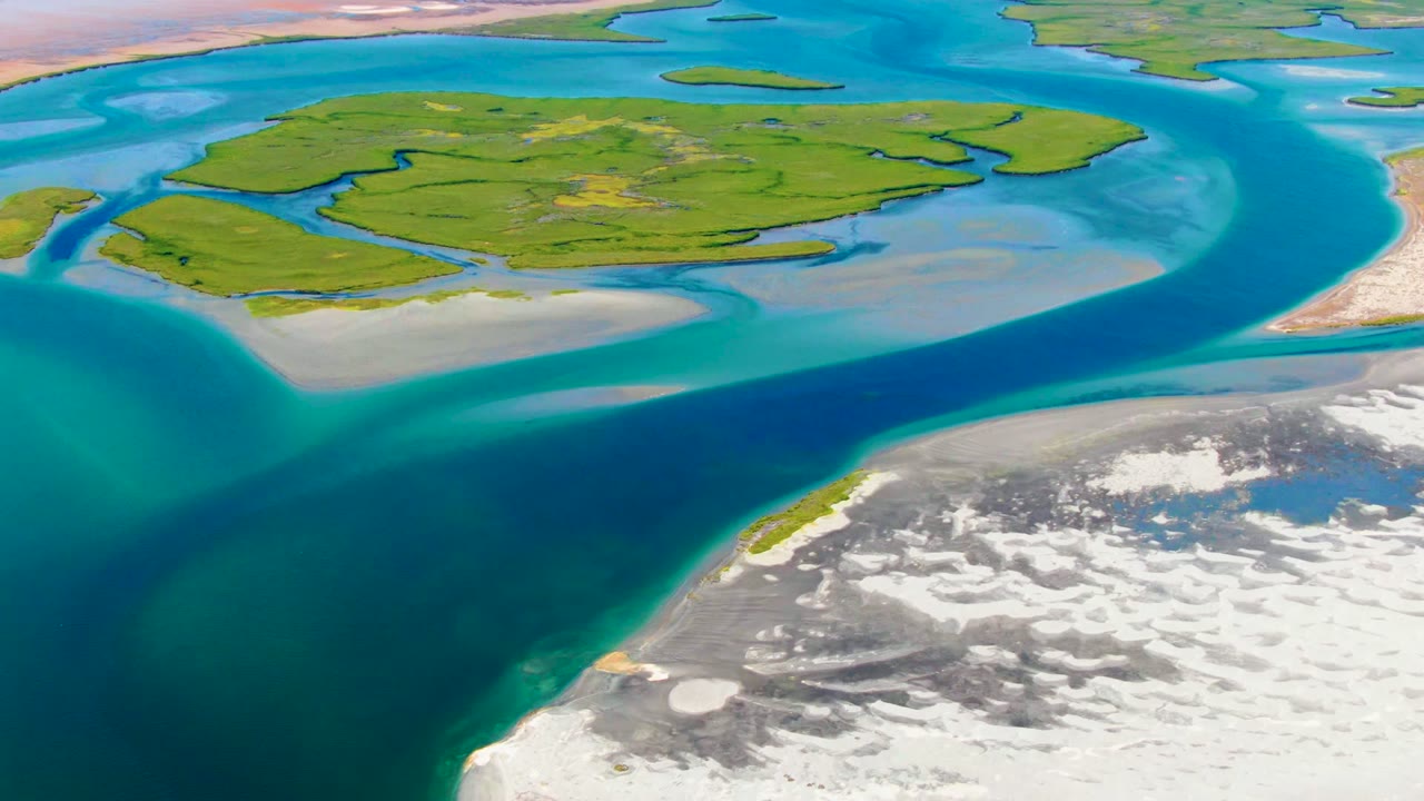 High aerial view of a green island, turquoise shallows, and a darker blue channel beside a rocky shore.