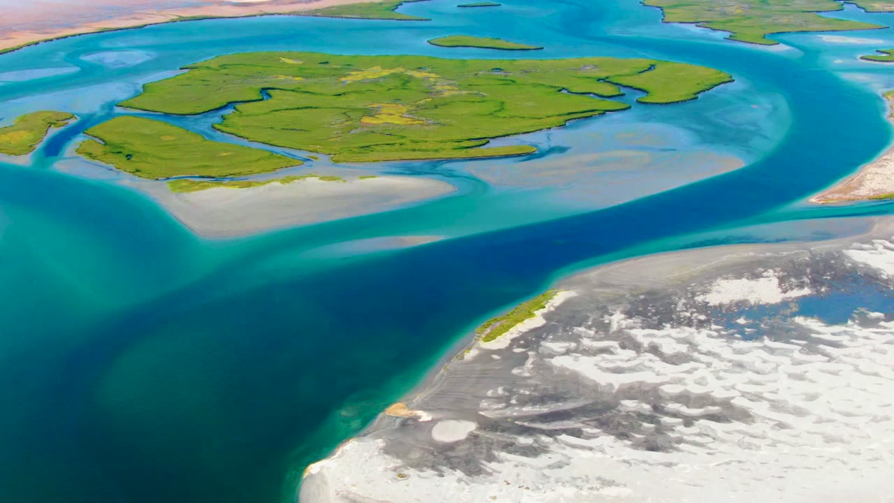 Aerial frame at 0:02 over La Bocana showing the green island across the upper frame, a broad blue channel through the center, pale shoreline edges, rocky foreground flats, and no visible people.
