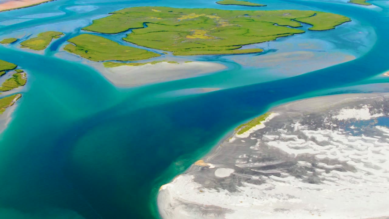 Aerial frame from La Bocana showing turquoise shallows, a darker central channel, green landmasses, and a bright sandbar along the water edge, no visible people.
