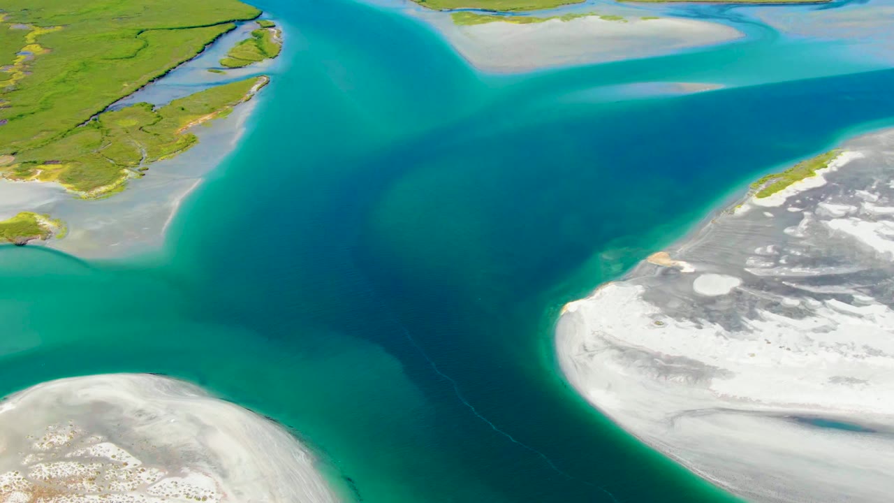 Aerial frame from La Bocana, Baja California Sur showing turquoise and deep blue water between white sandy shoals and green coastal land, with no visible people.