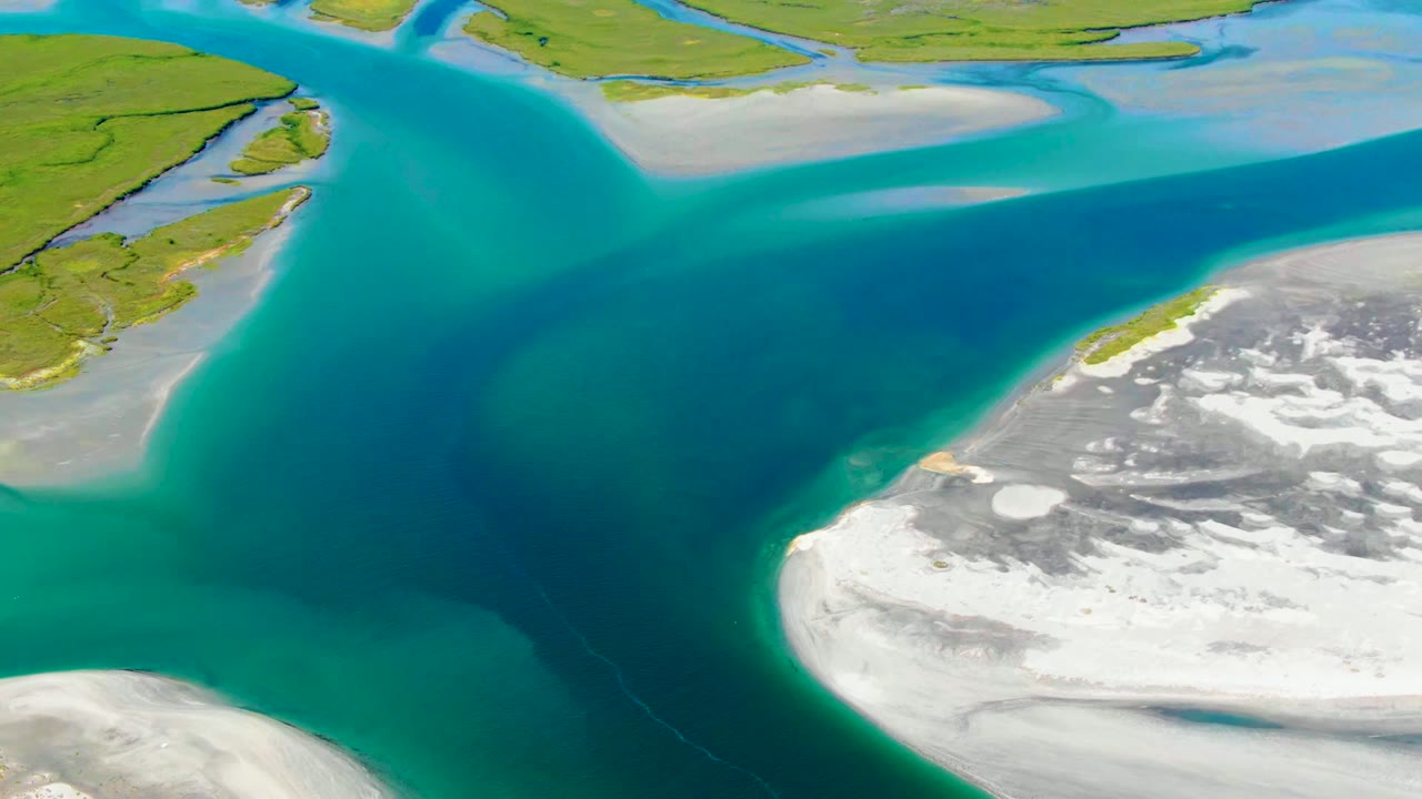 Aerial frame at 0:02 in La Bocana, Baja California Sur, with a curved turquoise channel, green land along the top, and white ice or snow along the lower right, no visible people.