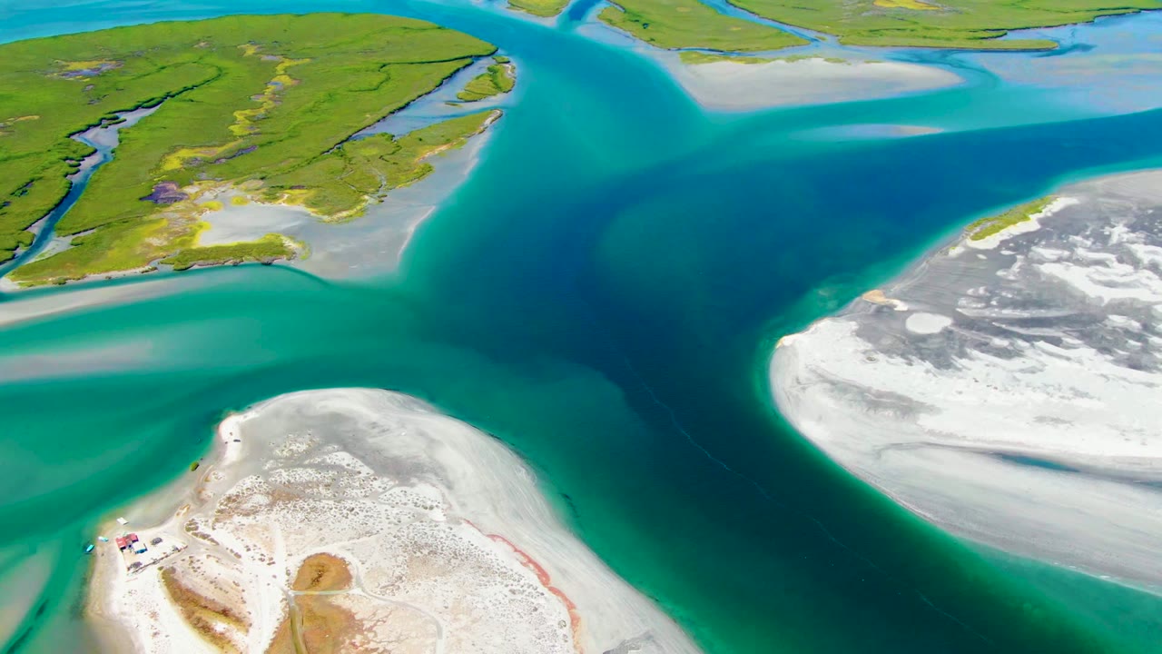 Drone view of a deep blue channel bordered by pale sand and green wetland islands.