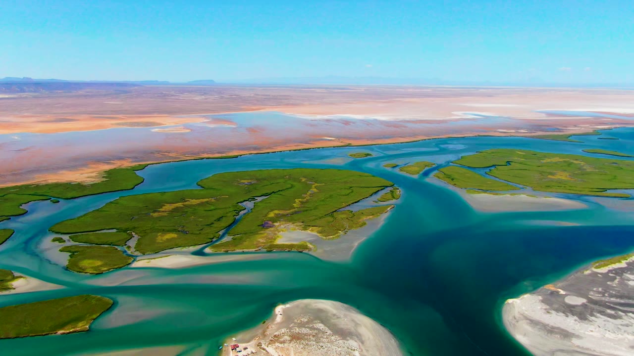 Aerial view over La Bocana, Baja California Sur, with turquoise water channels around green islands, pale sandbars in the foreground, desert flats on the horizon, and no visible people.
