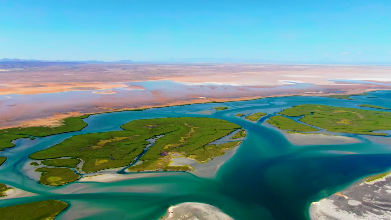 Opening aerial frame over La Bocana, Baja California Sur, with turquoise channels around green islands, pale sandbars at the bottom edge, a tan plain beyond, and no visible people.