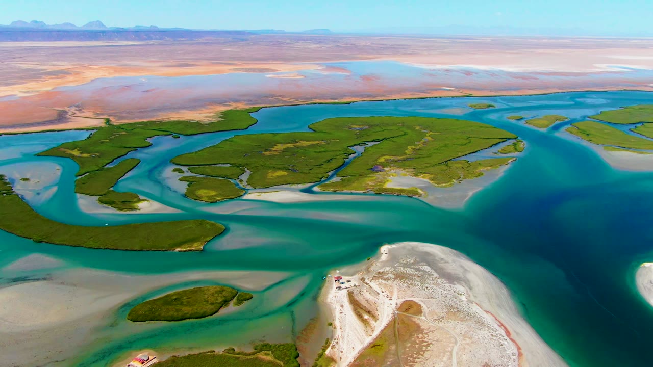 Aerial frame over La Bocana, Baja California Sur, with turquoise water channels around green delta islands, pale sandbars in the foreground, desert in the distance, and no visible people.