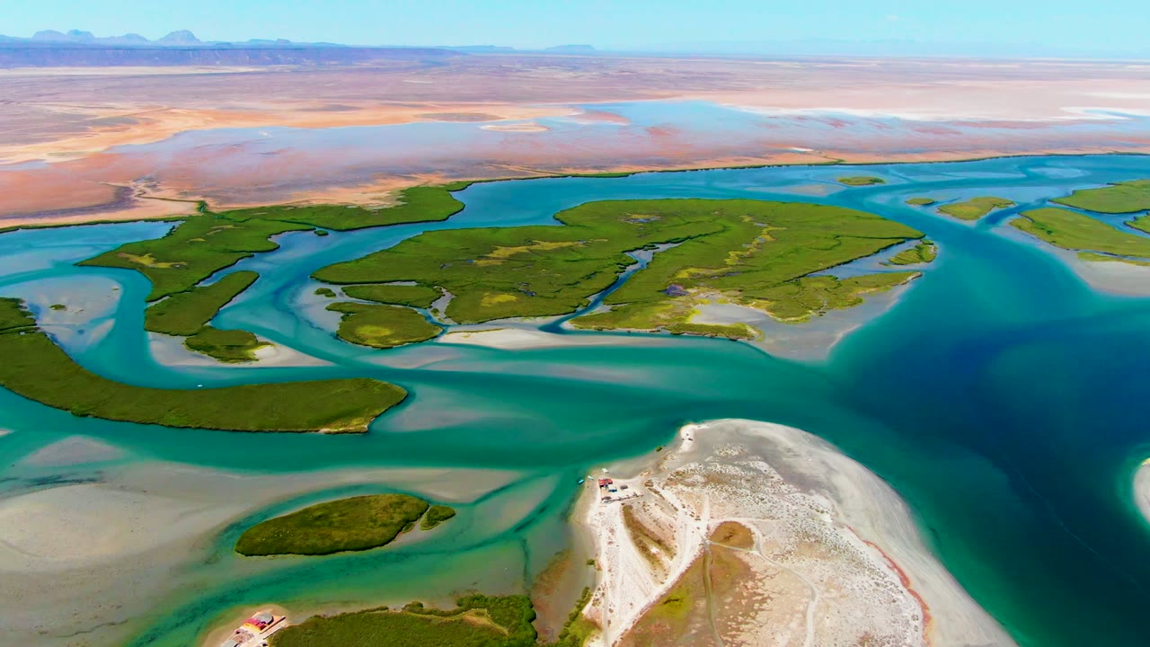 Drone view across green delta islands surrounded by blue water and pale sand.