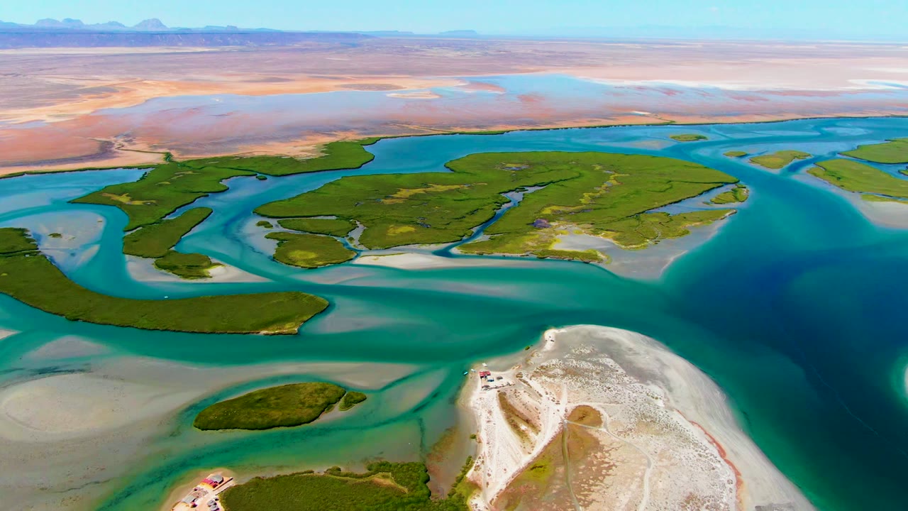 Aerial frame over La Bocana showing the broad delta from above, with a curved turquoise channel at left, larger green landmass in the center, pale sandbars, and no visible people.