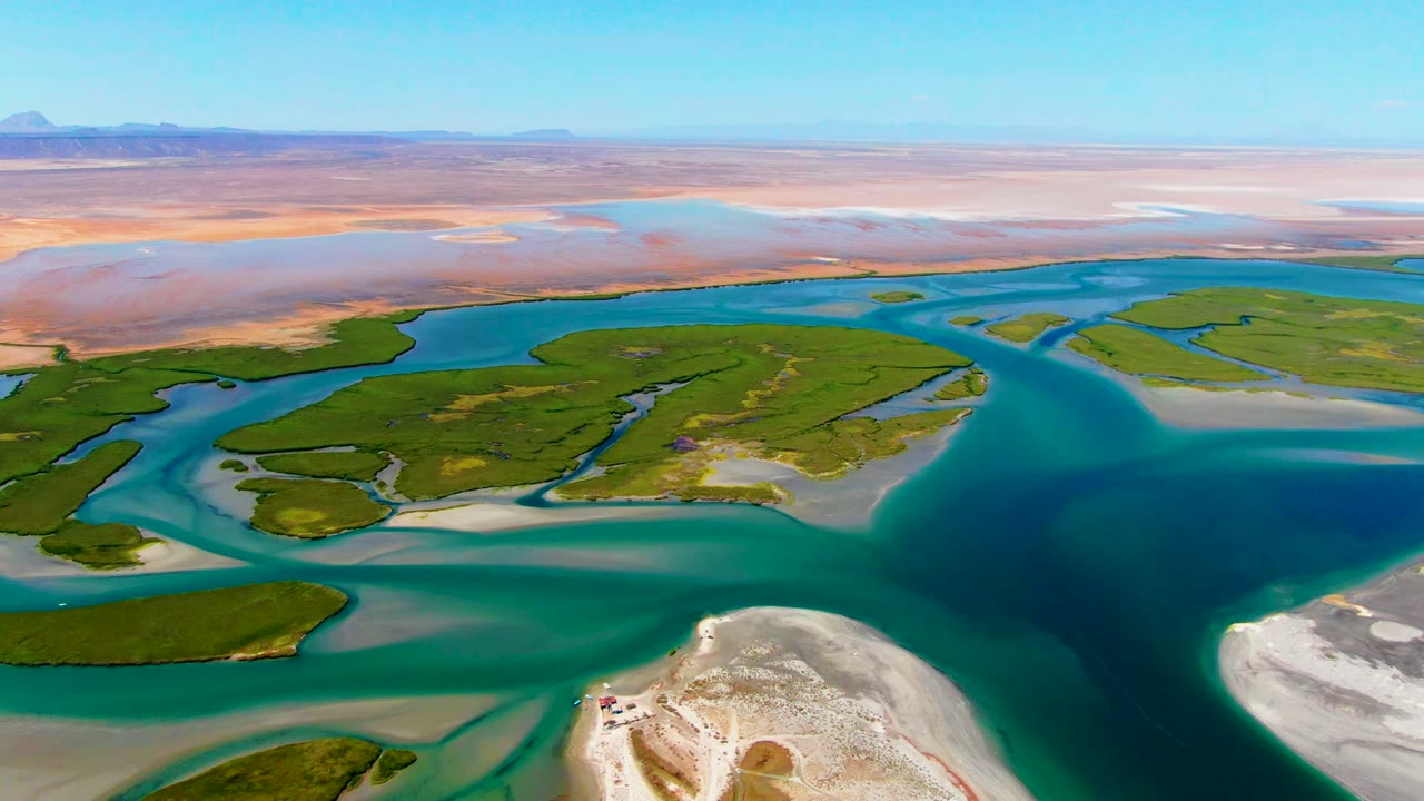 Aerial view over La Bocana, Baja California Sur, with turquoise channels around green islands, pale foreground sandbars, distant desert flats, and no visible people.