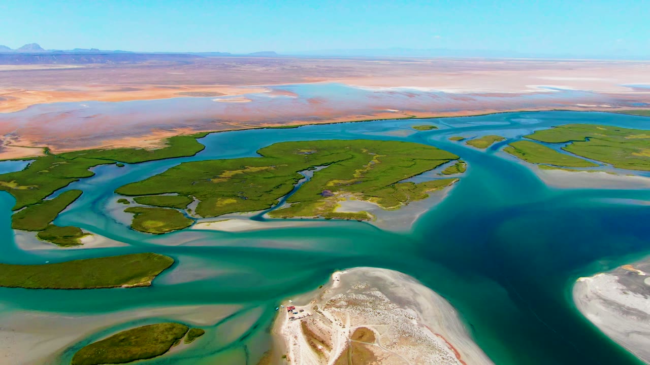 Drone perspective over La Bocana with a sandy foreground point, blue-green channels curving through low islands, dry desert beyond, and no visible people.