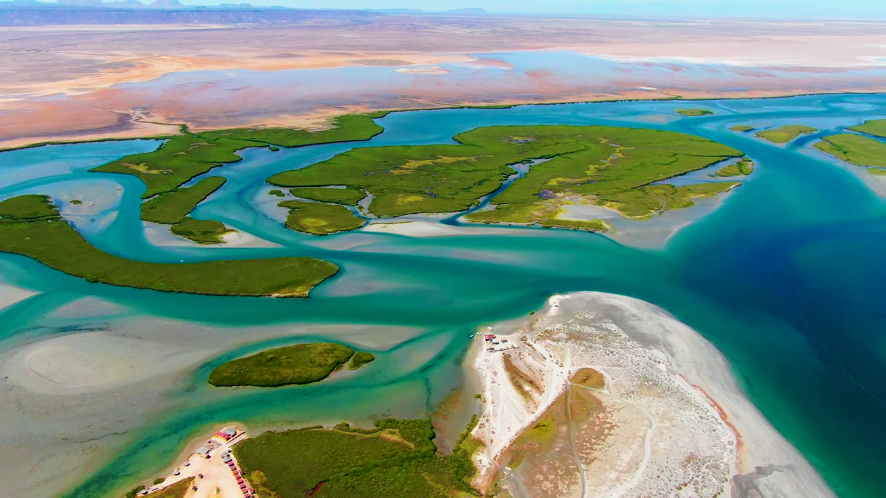 Aerial frame from La Bocana, Baja California Sur, showing green coastal islands, turquoise water channels, pale sandbars, a sandy foreground shore, and no visible people.