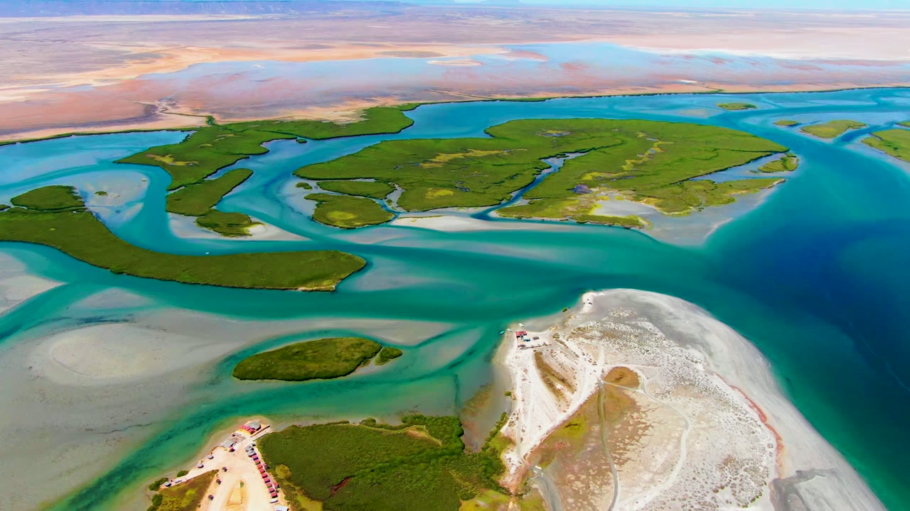 La Bocana aerial frame with blue-green water curving around low green islands, exposed pale sandbars on the left, a bright sandy shoreline in the foreground, and no visible people.