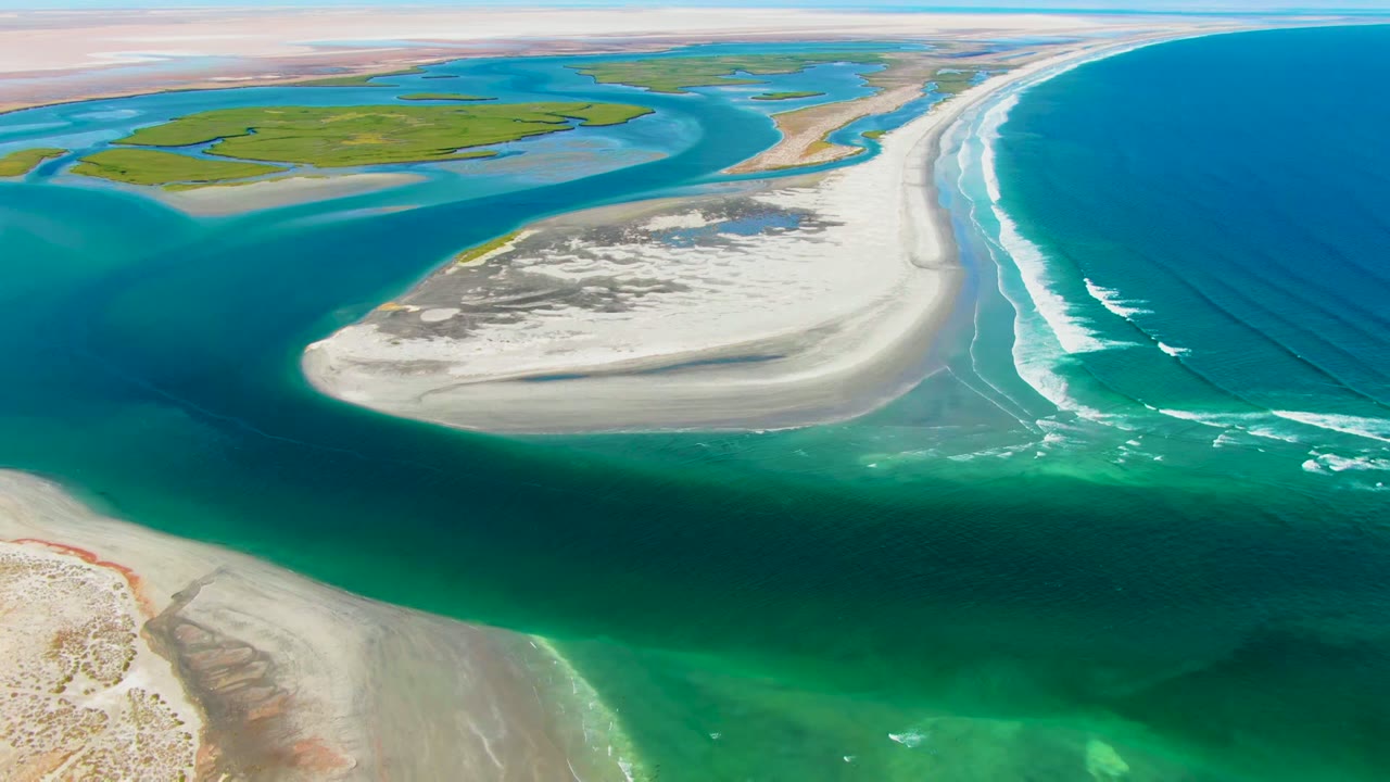Aerial frame from La Bocana, Baja California Sur showing a pale sandbar between turquoise estuary water and deep blue ocean surf, with green wetlands in the background and no visible people.