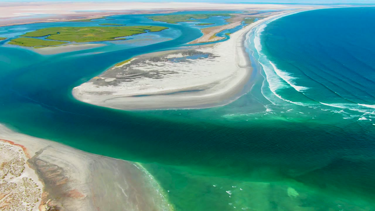 Aerial frame of a pale sandbar at La Bocana, Baja California Sur, with green shallows in front, white surf to the right, lagoon channels behind, and no visible people.