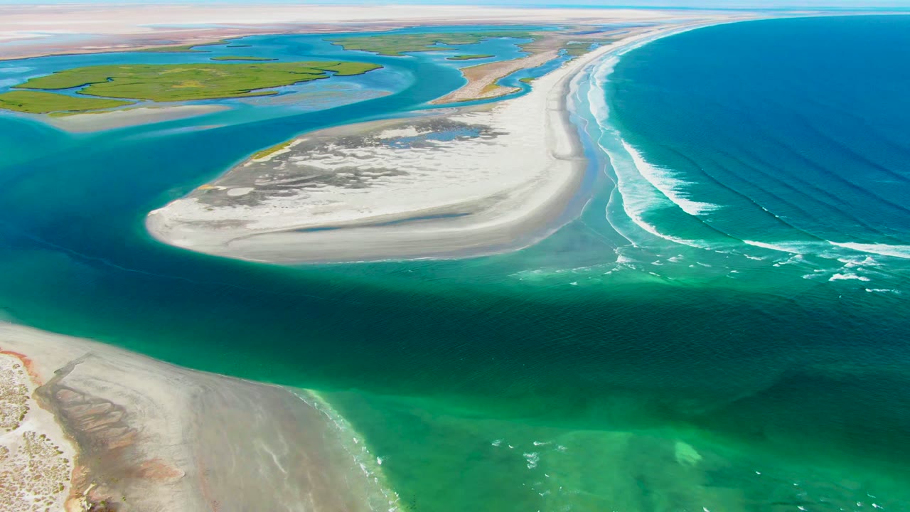 Wide La Bocana aerial frame with the sandbar centered, deeper blue ocean on the right, green shallow water below, low coastal land in the background, and no visible people.