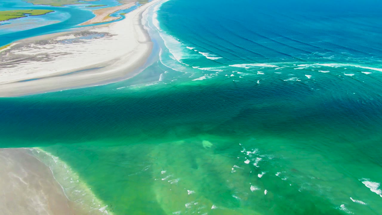 Aerial frame from La Bocana showing a broad blue ocean surface, curved pale sandbars, turquoise nearshore water, and scattered white surf with no visible people.