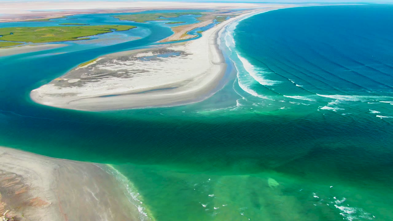 Wide aerial coastal sandbar with shallow green water in the foreground and surf offshore.