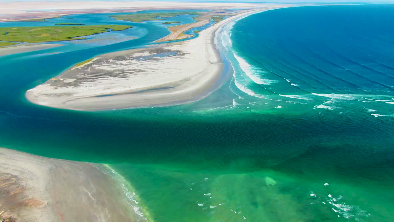 Aerial frame from La Bocana, Baja California Sur showing the sandbar centered below green lagoon channels and beside blue ocean surf, with no visible people.